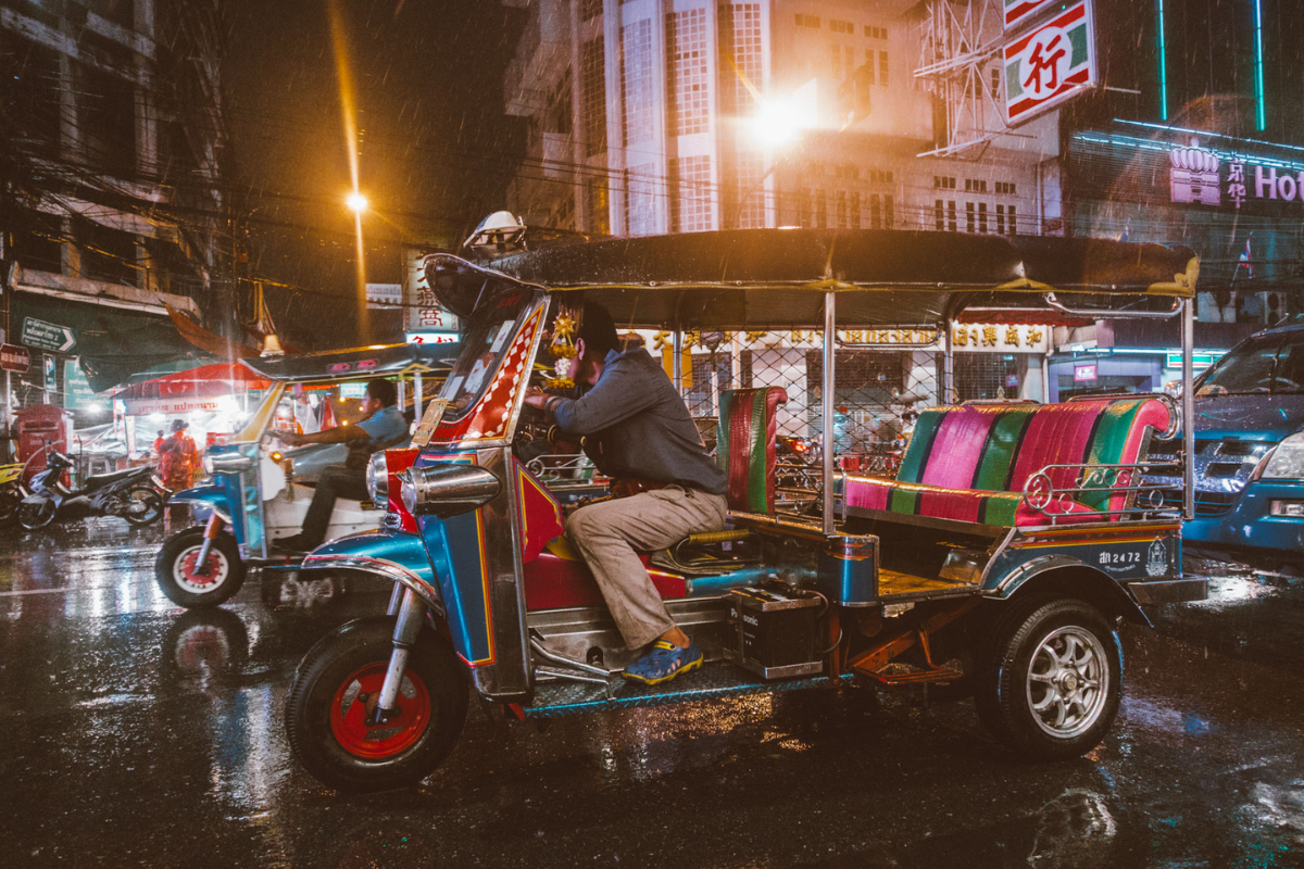 Rainy street scene in Bangkok with tuk-tuks and pedestrians under umbrellas